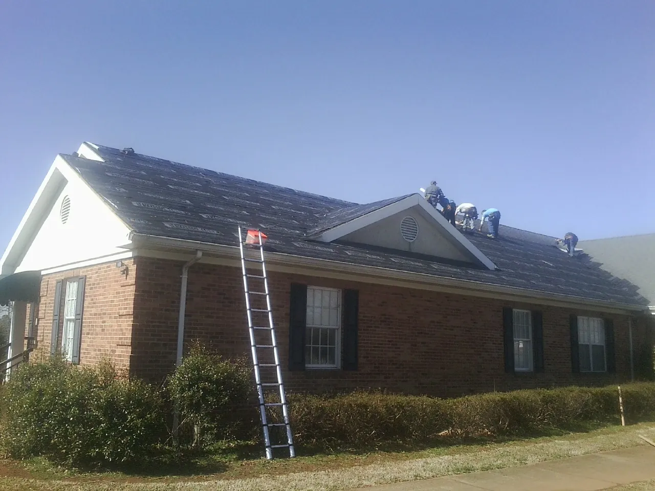 Crew tearing shingles off a brick ranch roof with a ladder set — Metal Roof Repair work by our crew in Bayville