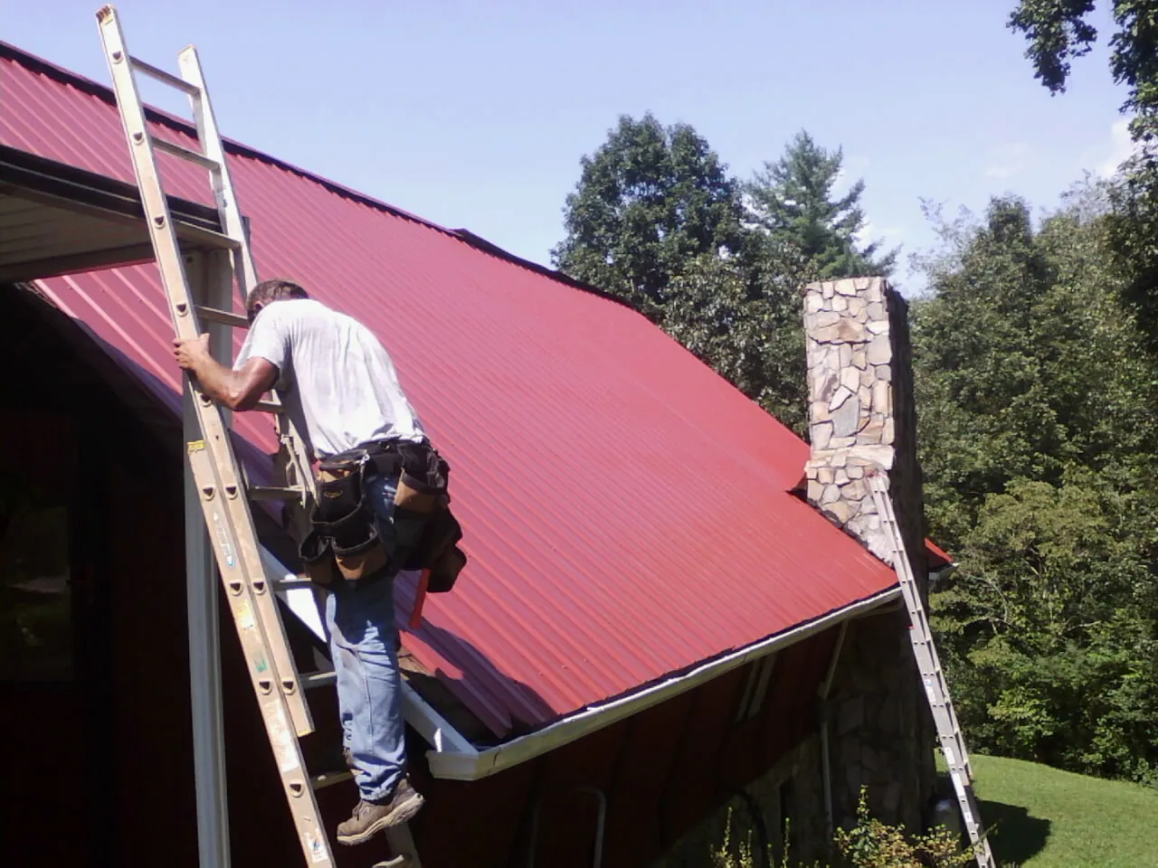 Roofer on a ladder beside a red metal roof and stone chimney — Soffit & Fascia Repair by Roofing Brothers in Bayville